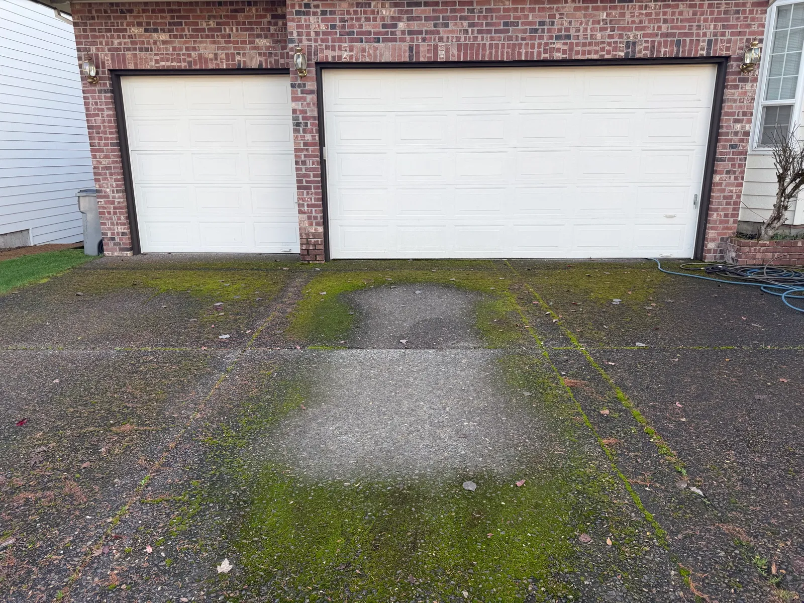 A clean test patch cut through a moss-covered driveway in front of a brick house