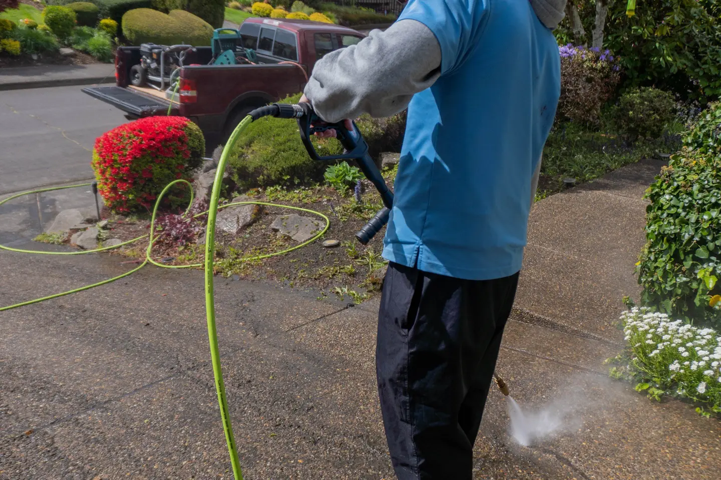 Pressure washing a driveway: wand and green hose in hand, mist on the concrete, red azaleas and the work truck behind