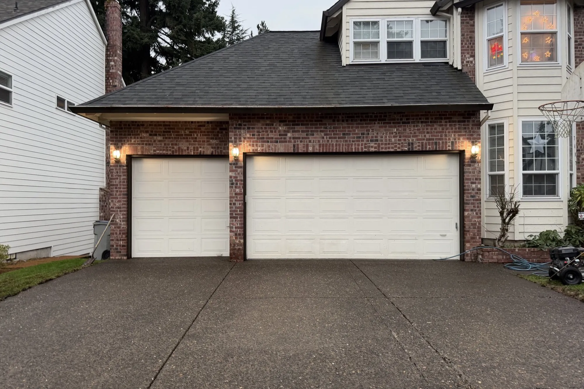 Clean two-car concrete driveway in front of a brick garage at dusk, porch lights on, tall firs behind the house